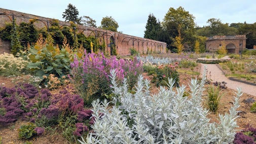 Newly emerging flowers in flower beds with accessible paths, old red brick wall and Triumphal Arch in the background
